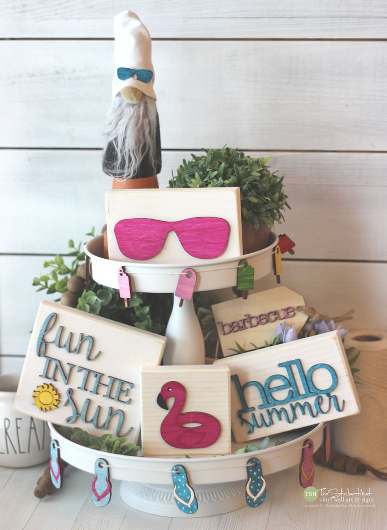 May include: A tiered tray decorated with summer-themed wooden signs. The signs feature phrases like "fun in the sun", "hello summer", and "barbecue". There are also pink sunglasses, a pink flamingo, and flip-flops.
