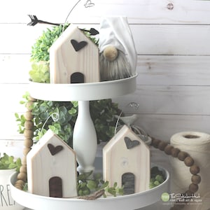 May include: Three wooden houses with heart accents on a tiered white stand. The houses are painted white with brown and gray accents. The stand is decorated with greenery and wooden beads.
