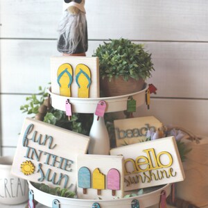 May include: A tiered tray decorated with summer-themed wooden signs. The signs feature phrases like "fun in the sun", "hello sunshine", and "beach". There are also wooden cutouts of flip flops and popsicles.
