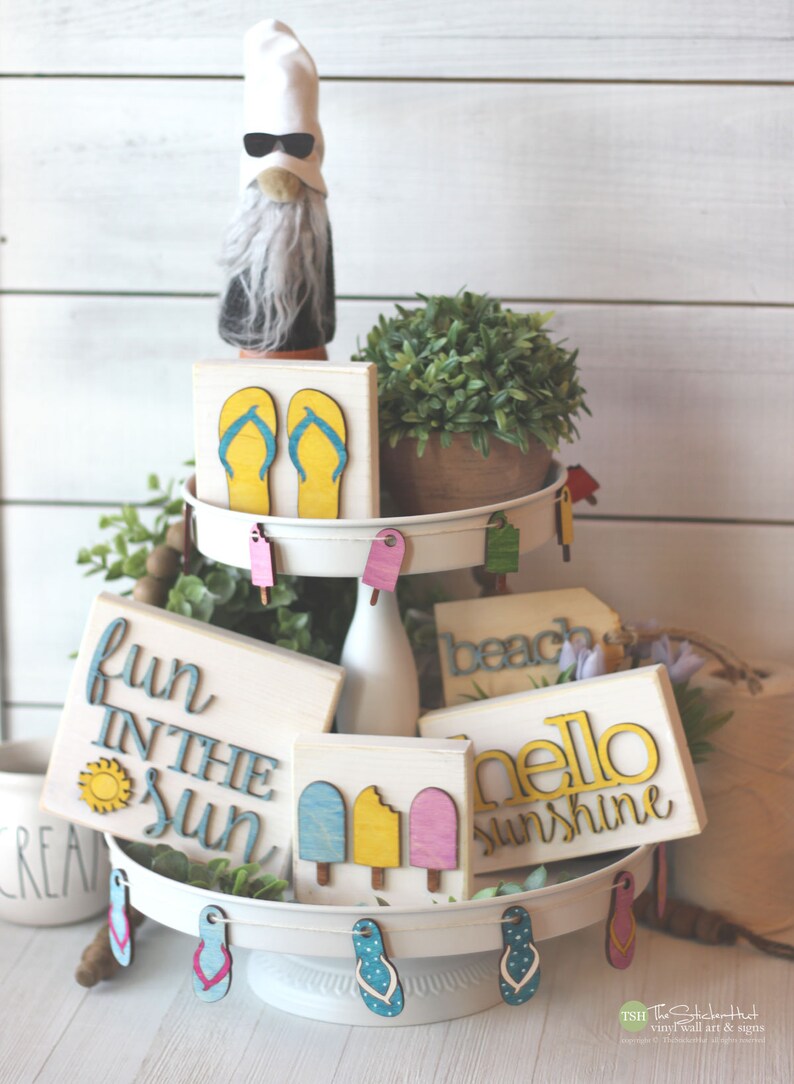 May include: A tiered tray decorated with summer-themed wooden signs. The signs feature phrases like "fun in the sun", "hello sunshine", and "beach". There are also wooden cutouts of flip flops and popsicles.