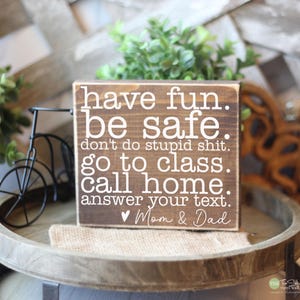 May include: A rustic wooden block sign with white lettering that reads "have fun. be safe. don't do stupid shit. go to class. call home. answer your text. Mom & Dad." The sign is on a wooden tray with decorative items.