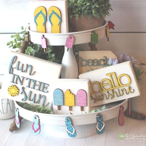 May include: A tiered tray decorated with wooden signs featuring summer-themed phrases like "fun in the sun", "beach", and "hello sunshine". The signs are painted in bright colors and have a rustic, handmade look. The tray is also decorated with wooden flip-flop cutouts.