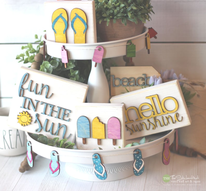 May include: A tiered tray decorated with wooden signs featuring summer-themed phrases like "fun in the sun", "beach", and "hello sunshine". The signs are painted in bright colors and have a rustic, handmade look. The tray is also decorated with wooden flip-flop cutouts.