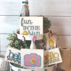 May include: A tiered tray decorated with summer-themed wooden signs. The top tier features a sign that says "fun in the sun" with a yellow sun graphic. The middle tier has a sign that says "hello sunshine" and a sign that says "hello summer". The bottom tier has a sign with a camera graphic and a heart.