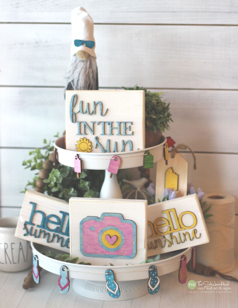 May include: A tiered tray decorated with summer-themed wooden signs. The top tier features a sign that says "fun in the sun" with a yellow sun graphic. The middle tier has a sign that says "hello sunshine" and a sign that says "hello summer". The bottom tier has a sign with a camera graphic and a heart.