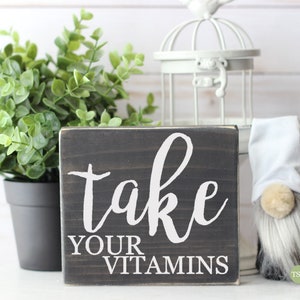 May include: A small wooden sign with the text "take your vitamins" in white paint on a black background. The sign is sitting on a white surface with a small potted plant and a white birdcage in the background.