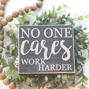 May include: A black wooden sign with white text that reads "No one cares work harder". The sign is sitting on a white surface with green plants and brown wooden beads.
