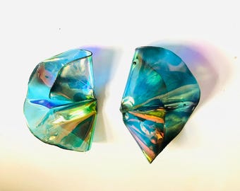 May include: Two iridescent, abstract-shaped objects with a folded design. The objects have a color palette of blues, greens, and hints of orange. They are set against a white background, creating a striking contrast.