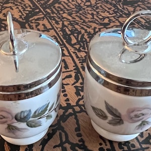 May include: Two white ceramic lidded jars featuring a floral pattern of soft pink roses and green leaves. Each jar has a polished silver-tone metal lid with a circular handle on top and a matching silver-tone band around the upper rim.