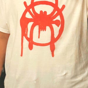 K&ouml;nnte beinhalten: Wei&szlig;es T-Shirt mit roter Spinnen-Grafik. Das Design ist eine stilisierte Spinne mit einem kreisf&ouml;rmigen, netzartigen Umriss und tropfenden Akzenten. Das Shirt besteht aus einem weichen, hellen Stoff.