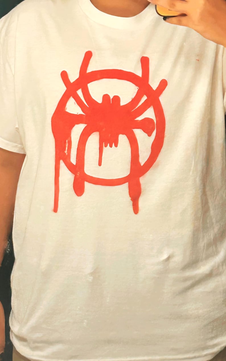 K&ouml;nnte beinhalten: Wei&szlig;es T-Shirt mit roter Spinnen-Grafik. Das Design ist eine stilisierte Spinne mit einem kreisf&ouml;rmigen, netzartigen Umriss und tropfenden Akzenten. Das Shirt besteht aus einem weichen, hellen Stoff.