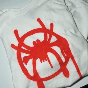 K&ouml;nnte beinhalten: Wei&szlig;es T-Shirt mit einem gro&szlig;en roten Spinnennetz-Design auf der Vorderseite. Die Grafik ist ein stilisiertes Spinnennetz im Spr&uuml;hstil. Das Shirt ist gefaltet und zeigt den Kragen und einen Teil des &Auml;rmels.