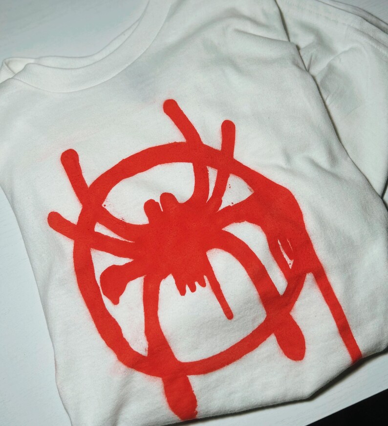 K&ouml;nnte beinhalten: Wei&szlig;es T-Shirt mit einem gro&szlig;en roten Spinnennetz-Design auf der Vorderseite. Die Grafik ist ein stilisiertes Spinnennetz im Spr&uuml;hstil. Das Shirt ist gefaltet und zeigt den Kragen und einen Teil des &Auml;rmels.