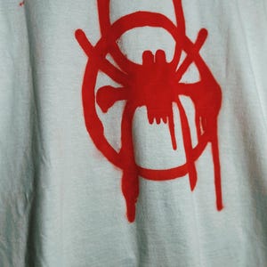 K&ouml;nnte beinhalten: Wei&szlig;es T-Shirt mit roter Spinnengrafik in einem Kreis, mit tropfendem Farbeffekt. Das Design ist mittig auf dem T-Shirt platziert. Der Stoff wirkt leicht zerknittert.