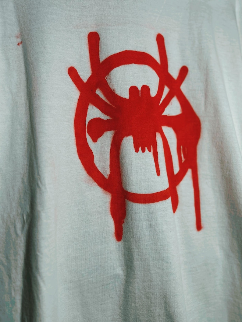 K&ouml;nnte beinhalten: Wei&szlig;es T-Shirt mit roter Spinnengrafik in einem Kreis, mit tropfendem Farbeffekt. Das Design ist mittig auf dem T-Shirt platziert. Der Stoff wirkt leicht zerknittert.