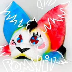 May include: A colorful, plush animal mask with rainbow-colored ears and accents. The mask has large, expressive eyes with red and blue details, a black nose, and the word "POMMII!" written in white.