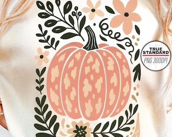 Może przedstawiać: Kremowy t-shirt z brzoskwiniowym wzorem dyni otoczonym zielonymi liśćmi i brzoskwiniowymi kwiatami. Dynia jest centralnym elementem. Tekst "TRUE STANDARD PNG 3000PI" znajduje się w rogu.