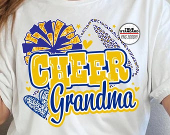 Puede incluir: Camiseta blanca con las palabras "CHEER Grandma" en amarillo y azul. El diseño incluye un pompón, corazones y un borde con estampado de leopardo. La camiseta es de estilo casual.