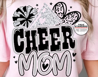 Może przedstawiać: Różowa koszulka z czarno-białym nadrukiem, na którym widnieje napis "Cheer Mom" z efektem srebrnego brokatu. Nadruk zawiera również czarno-biały pompon cheerleaderski, srebrną błyszczącą gwiazdę i czarno-biały kwiat w panterkę.