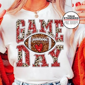 May include: White t-shirt with a red, brown, and gold glitter design that says "GAME DAY" with a football and heart in the center.