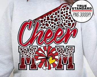 Puede incluir: Una sudadera gris con un diseño de porrista rojo y blanco. El diseño incluye un megáfono rojo con estampado de leopardo, una pompon rojo y blanco y las palabras "Cheer Mom" en purpurina roja. El diseño está rodeado por un borde de purpurina roja.