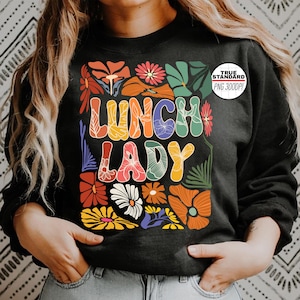 Puede incluir: Sudadera negra con un diseño floral colorido y las palabras "LUNCH LADY" en una fuente retro. El diseño incluye flores naranjas, amarillas, rojas y azules y hojas verdes. Un círculo blanco con "TRUE STANDARD PNG 300DPI" está en la esquina superior derecha.