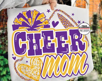 Puede incluir: Camiseta blanca con un diseño morado y dorado. El diseño incluye las palabras "CHEER MOM" en negrita, un pompón de animadora, corazones y estrellas. El diseño tiene un estampado de leopardo.
