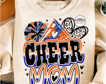 Może przedstawiać: Kremowa bluza z nadrukiem graficznym z napisem "CHEER MOM" pogrubioną, obrysowaną czcionką. Projekt zawiera pompon, serce i kształt domu z sercem, wszystko w kolorze niebieskim i pomarańczowym. Bluza jest złożona.