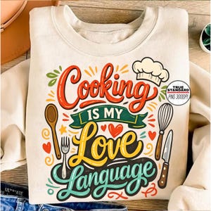 May include: Cream-colored sweatshirt with the text "Cooking Is My Love Language" in a colorful, whimsical design. The graphic includes a chef's hat, spoon, fork, whisk, knife, and hearts, perfect for anyone who loves to cook.