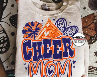Może przedstawiać: Beżowa bluza z żywym wzorem z napisem "CHEER MOM" w kolorze niebieskim i pomarańczowym. Wzór zawiera pompony, serca i błyszczący pomarańczowy trójkąt. Bluza jest złożona.