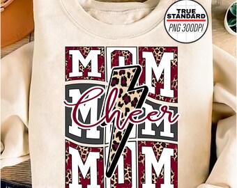 Puede incluir: Una sudadera blanca con un diseño gráfico rojo, negro y blanco que dice "Mom Cheer Mom" con un rayo de impresión de leopardo en el centro.