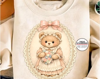 Puede incluir: Sudadera de color crema con una ilustración detallada de un oso de peluche con gorro y vestido, sosteniendo un edredón de retazos. El oso está enmarcado dentro de un óvalo decorativo con un lazo rosa en la parte superior. El texto "TRUE STANDARD PNG 300DPI" es visible.