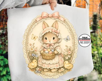 Puede incluir: Un bolso tote blanco con una ilustración vintage de un conejito de Pascua con gorro y vestido, sosteniendo una cesta de huevos. El diseño incluye mariposas, flores y dos pollitos, enmarcado por un borde ovalado de encaje.