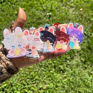 May include: A set of five clear acrylic charms featuring chibi-style characters with animal ears. Each charm displays a unique character with distinct hair colors including white, black, red, and purple, accented with small colorful star and heart details.