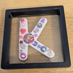 Puede incluir: Una letra K enmarcada decorada con botones, cuentas y estrellas de color rosa, morado y blanco.