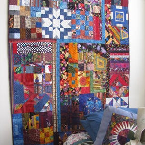 May include: A large, colorful quilt with a star pattern and various patchwork designs. The quilt features a mix of fabrics in shades of blue, red, orange, and green. The quilt is hanging on a wall, showcasing its intricate details and craftsmanship.
