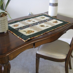 May include: A rectangular table runner with a green border and a patchwork design featuring images of gourds and sunflowers. The runner is placed on a dark brown wooden table, next to a decorative vase and a potted plant.