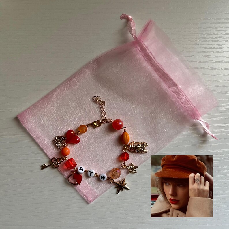 May include: A gold-toned charm bracelet with red, orange, and clear beads, and charms including a key, heart, star, and flower. The bracelet has the letters "ATW" on it. The bracelet is next to a pink organza bag.