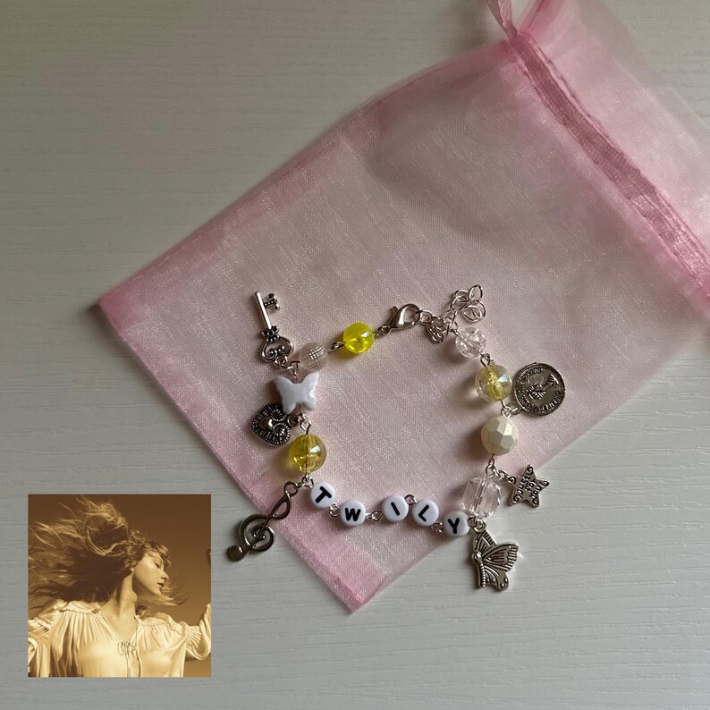 May include: A beaded bracelet with charms, including a key, butterfly, heart, and star. The bracelet features yellow and clear beads, and the word "TWILY" spelled out with letter beads. A pink organza bag is in the background.