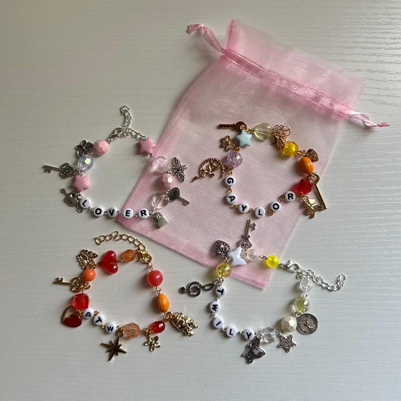 May include: Four beaded bracelets with charms and letter beads spelling out "LOVER", "TAYLOR", "ATW", and "TWILY". The bracelets feature keys, stars, and other decorative elements. A pink organza bag is in the background.