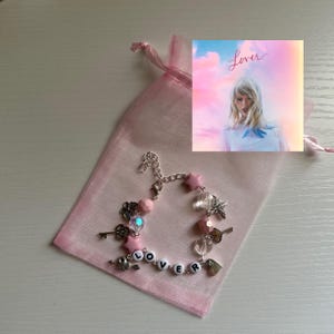 May include: A pink charm bracelet with star, key, and heart charms, spelling out "Lover" with letter beads. The bracelet is displayed in a pink organza bag, with a small image of the album cover.