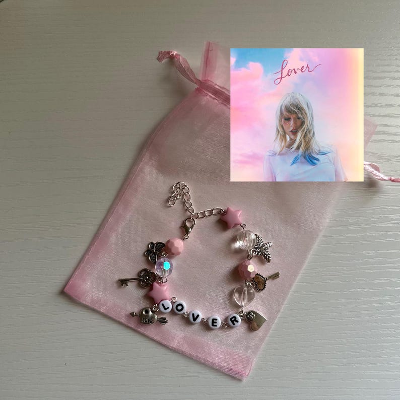 May include: A pink charm bracelet with star, key, and heart charms, spelling out "Lover" with letter beads. The bracelet is displayed in a pink organza bag, with a small image of the album cover.