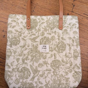 May include: A cream tote bag with a green floral design and brown leather handles. The bag has a rectangular shape and is adorned with a pattern of flowers and birds. The text "Elm Road Australia" is visible on a small label.