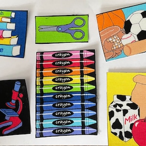 May include: A set of colorful felt patches with various school-themed images, including books, crayons, scissors, a microscope, a beaker, a lunch bag, a carton of milk, an apple, a baseball, a bat, a glove, and a soccer ball.