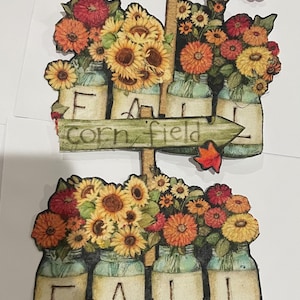 May include: Wooden sign with the words "SUNFLOWERS" and "corn field" pointing to mason jars filled with sunflowers and fall flowers. The word "FALL" is spelled out below in mason jars.