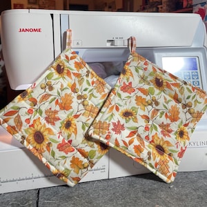May include: Two square fabric pot holders featuring a cream background with a colorful autumn print of sunflowers, orange and yellow oak leaves, and brown acorns. Each pot holder has a small checkered fabric loop for hanging at the top edge.