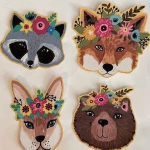 May include: Four colorful animal faces with floral crowns. A raccoon, fox, bunny, and bear with yellow outlines. The animals are all wearing flower crowns with pink, yellow, blue, and green flowers.