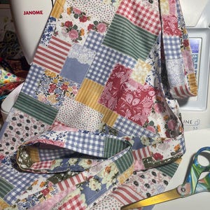 May include: A patchwork fabric project with a variety of floral, striped, and gingham patterns in red, pink, blue, and yellow. The fabric is draped over a white sewing machine. Gold scissors are visible.