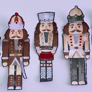 Puede incluir: Cinco adornos planos de madera con forma de cascanueces dispuestos en fila. Cada figura presenta uniformes pintados en tonos verde, marrón y crema, con detalles en rojo, adornos plateados y motivos de copos de nieve blancos en sombreros.