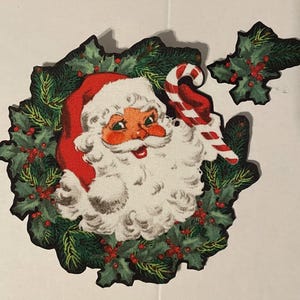 Puede incluir: Decoración navideña vintage con Papá Noel con gorro rojo y barba blanca, rodeado por una corona de acebo verde y bayas rojas. También se ve un bastón de caramelo. La decoración está recortada y tiene un diseño clásico y festivo.